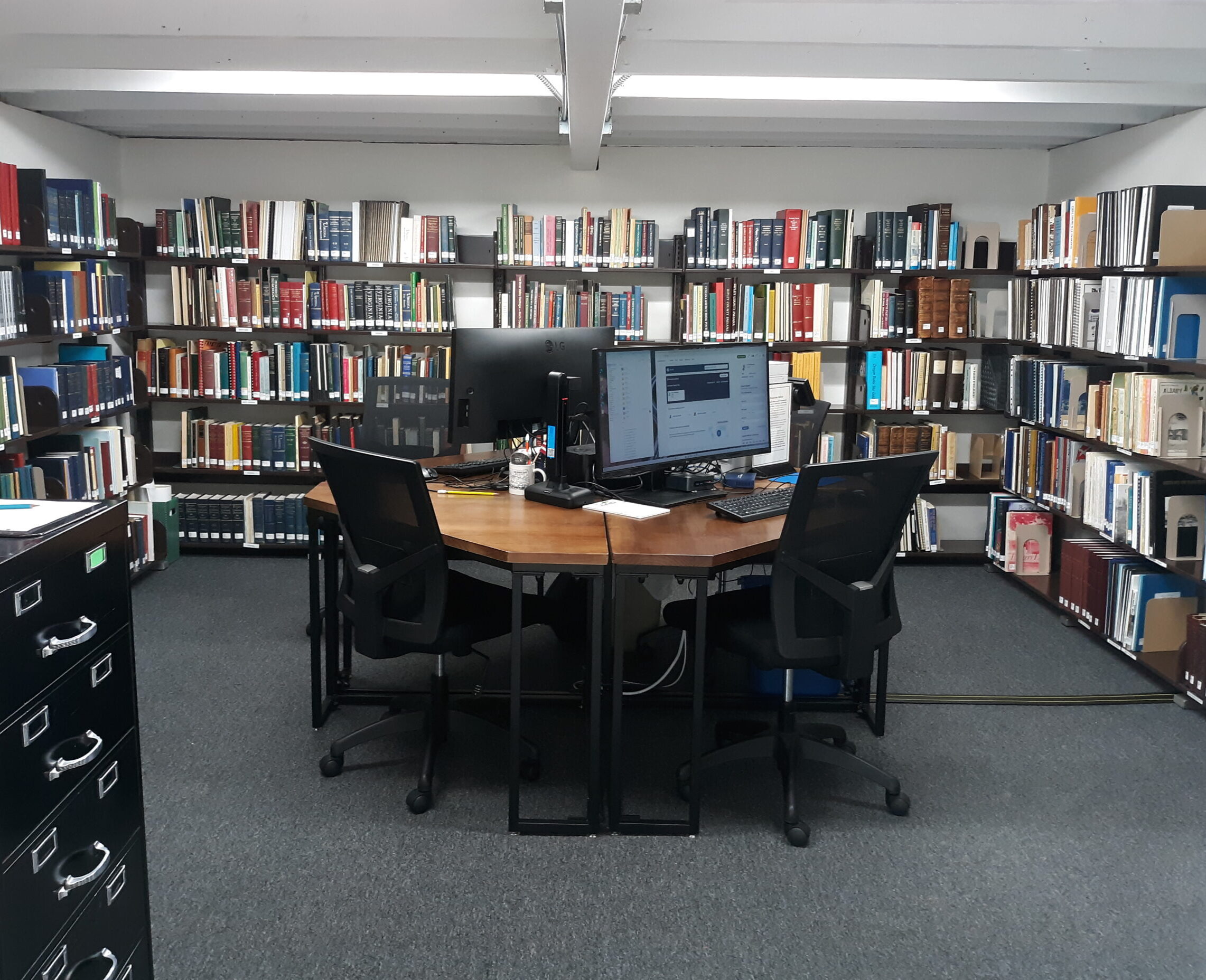 inside the library are two computer screens at a shared desk in the middle of a room lined with books and documents